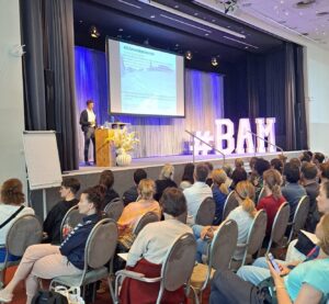 Bam Veranstaltung Berlin, Dr. Reto Schwenke auf der Bühne, mit einem großen BAM Dekostück. Publikum sitzen vor der Bühne und hören gespannt seinem Vortrag zu