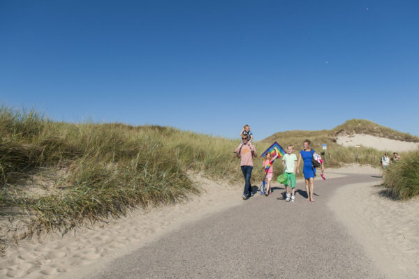 Düne mit Spazierweg und einer Familie. Papa hat Kind auf den Schultern und ein Kind hält einen bunten Drachsen in der Hand